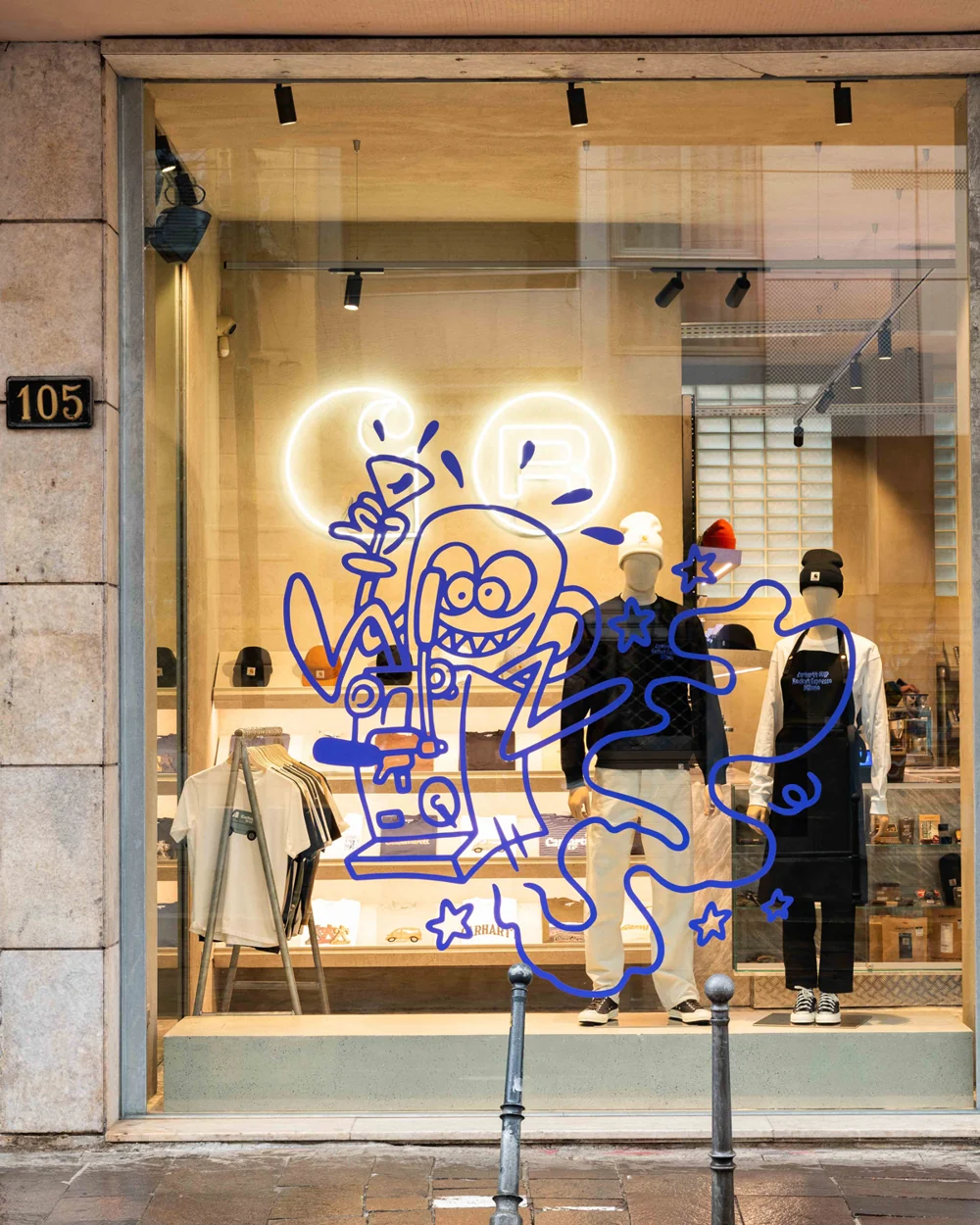 Blue collaboration artwork across the Milano Ticinese store window, with clothing displayed behind it